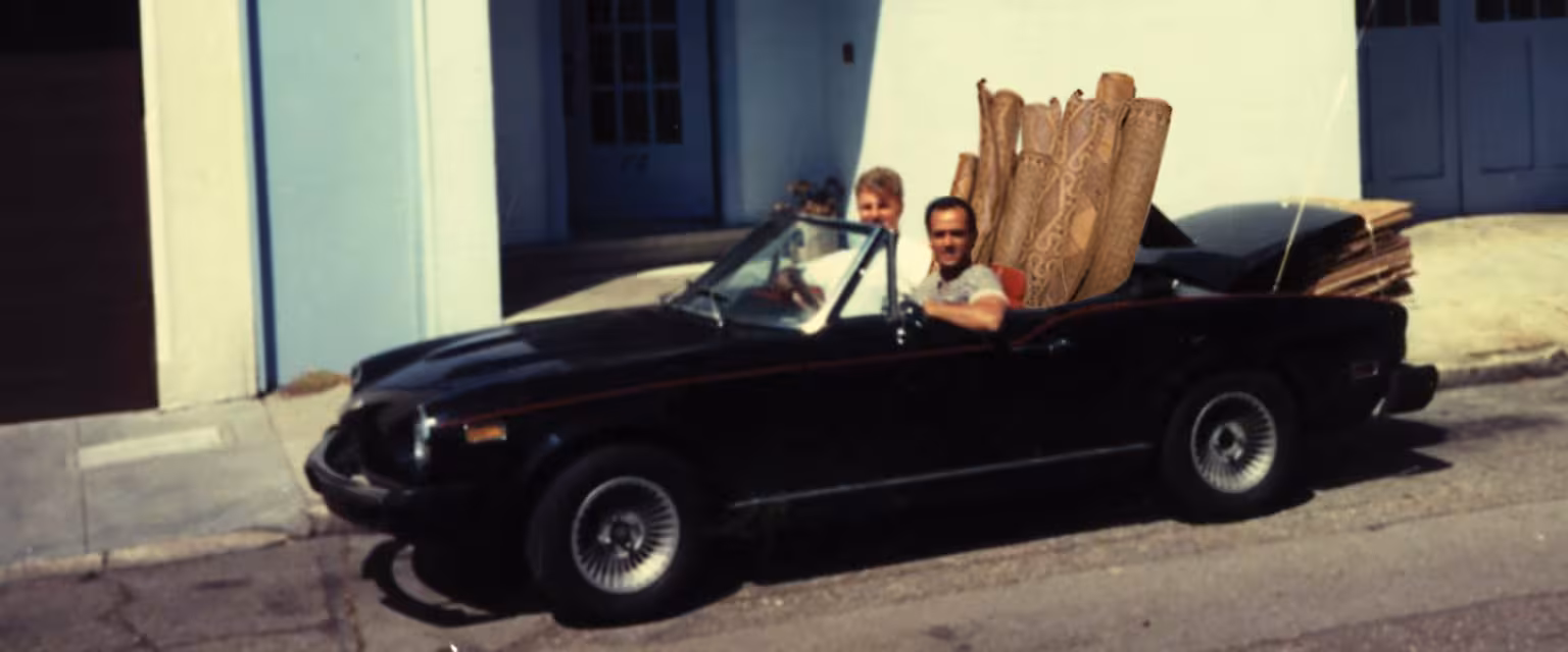 Two people driving a convertible car filled with wooden planks, parked beside a light blue building